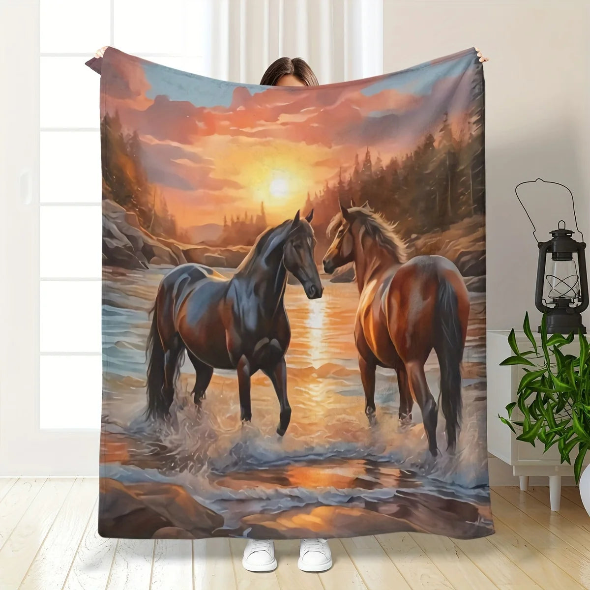 A person holds up a large fleece blanket featuring a scenic print of two horses standing in a river at sunset, with a forest and glowing sky in the background. The blanket is displayed in a bright indoor room with a window, floor, lantern, and plant visible.