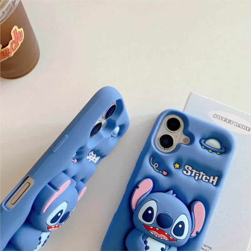 Two blue phone cases featuring a 3D cartoon character design resembling Stitch, shown on a light beige tabletop with a coffee cup and a small card in the background.