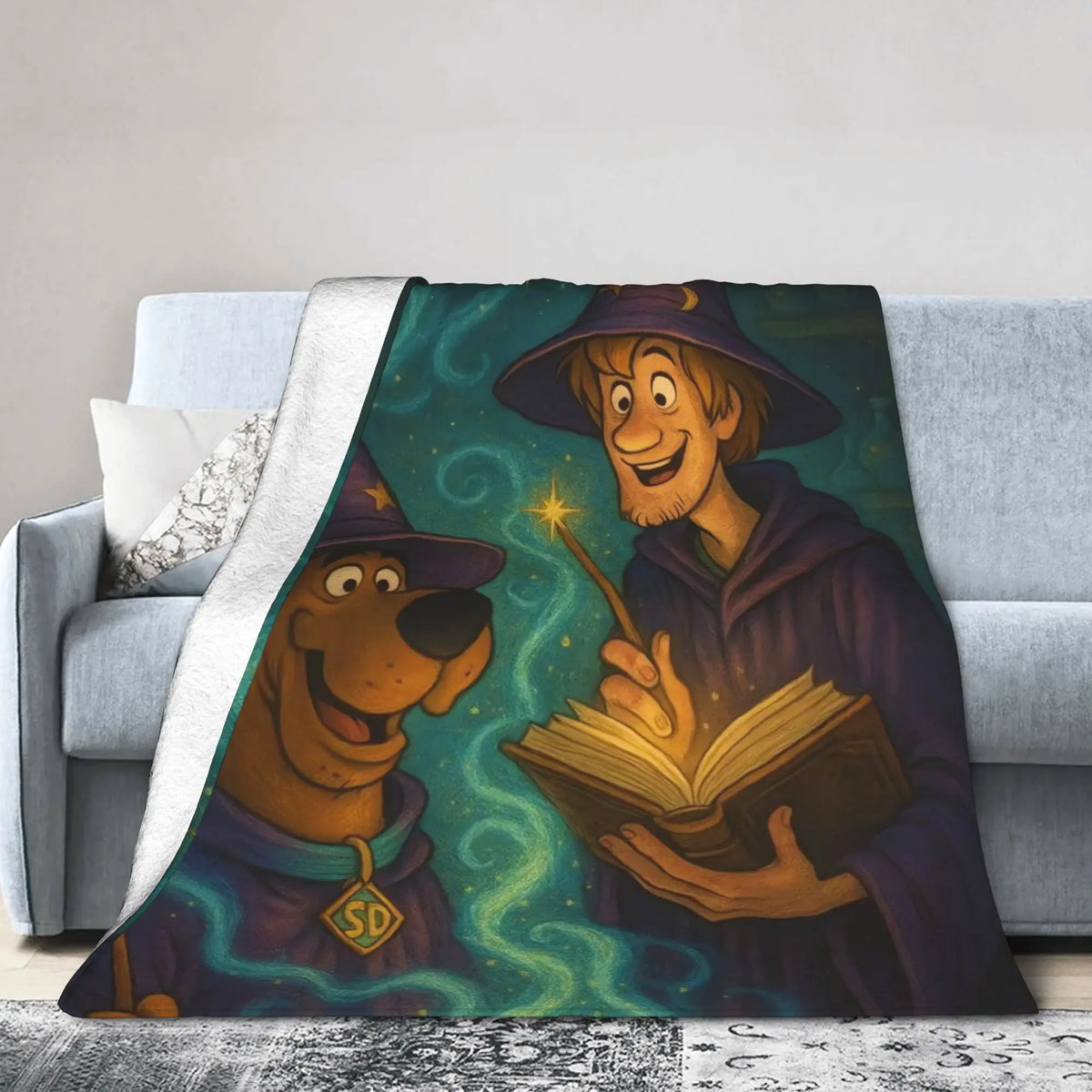 A fleece throw blanket draped over a light gray sofa, featuring a colorful cartoon illustration of Scooby-Doo and a wizard holding a glowing book. The blanket has a white sherpa backing.