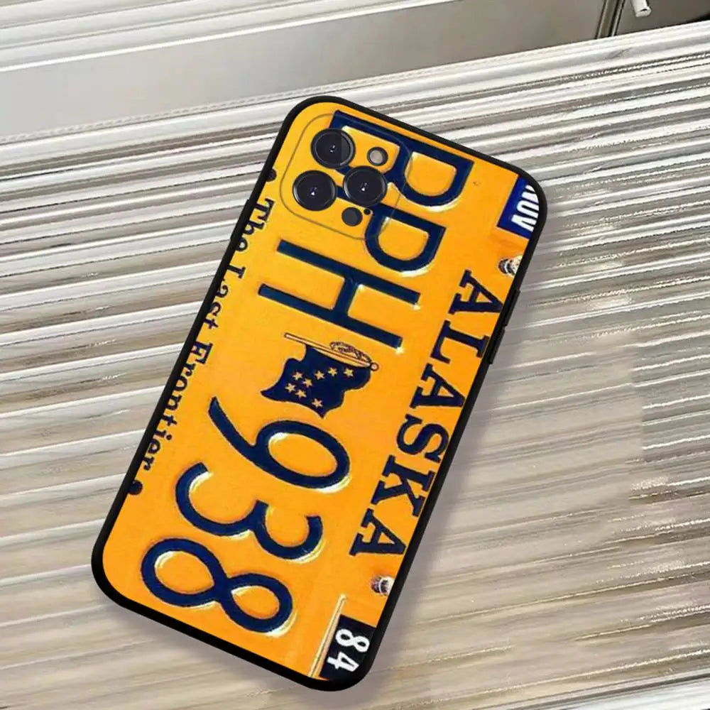 A phone case designed to look like an Alaska license plate, featuring a bright yellow background, bold blue lettering, and a black border, shown resting on a corrugated silver metal surface.