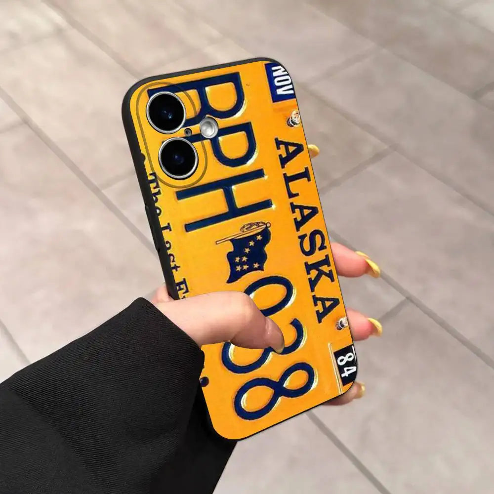 A hand holding a yellow phone case designed to look like an Alaska license plate, with the text 'ALASKA', 'BP 038', and 'NOW' in dark blue, set against a tiled floor background.