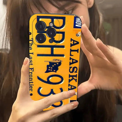 A yellow phone case designed like an Alaska license plate, held up by a person taking a mirror selfie. The case features dark blue text reading 'ALASKA', 'The Last Frontier', and '93', along with a small state flag graphic.