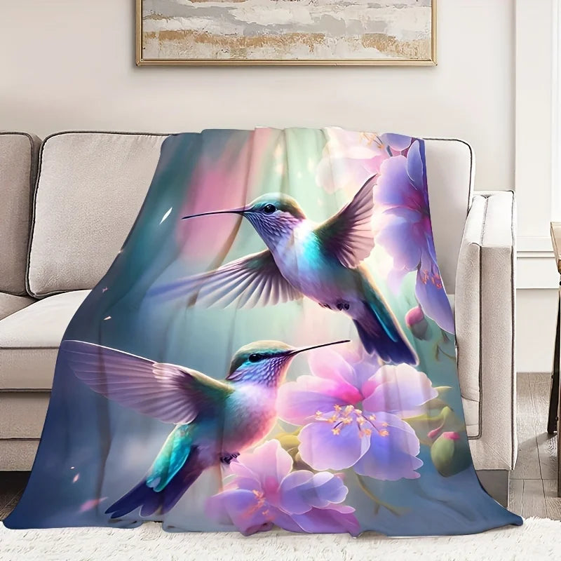 A soft throw blanket draped over a light beige sofa in a living room setting, featuring a colorful printed design of two hummingbirds and pink flowers.
