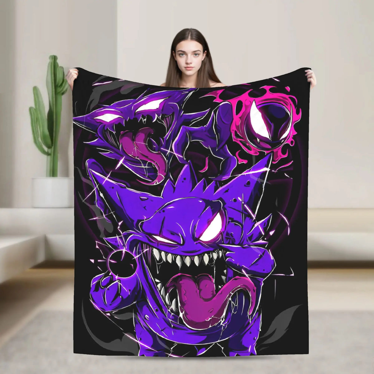 A woman holds up a large black blanket or throw featuring a bold purple and pink monster graphic with glowing white eyes, shown in a bright indoor room with a cactus in the background.