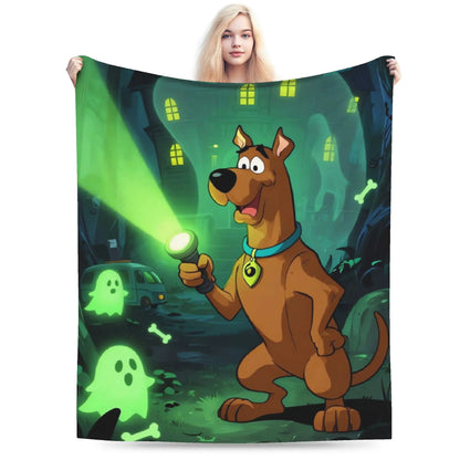 A person holds up a large printed blanket or tapestry featuring Scooby-Doo standing in a spooky haunted-house scene with a green flashlight beam, glowing ghosts, and bones. The product is shown against a plain white background, with the model's head partially visible above it.