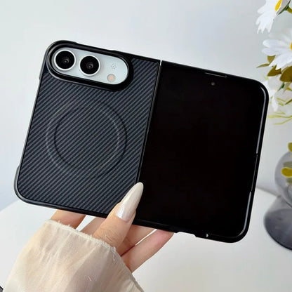 A hand with long nude manicured nails holds a black flip-style phone case with a carbon fiber textured back and a glossy black front cover, shown against a bright white background with a blurred flower in the corner.