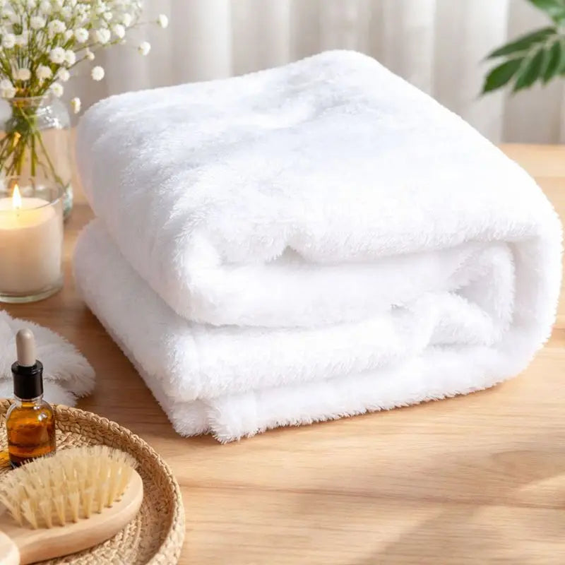 A stack of two fluffy white towels on a light wood surface, styled with spa-like props including a candle, small bottle, brush, and flowers in the background.