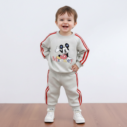 A smiling toddler stands on a wooden floor against a plain light background, wearing a light gray two-piece tracksuit with red side stripes and a Mickey Mouse graphic on the chest, plus white sneakers.