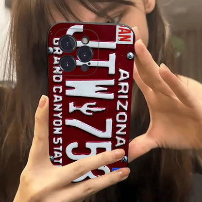 A close-up of a person holding a red Arizona license plate phone case in front of their face, showing the case design with white lettering and silver screws.