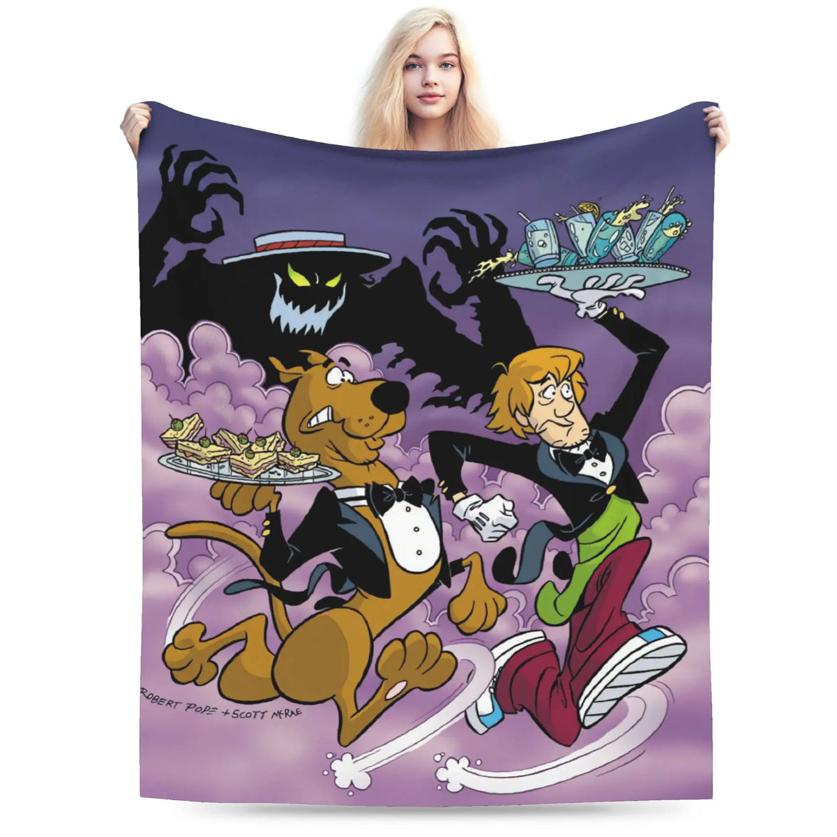 A person holds up a large square blanket or throw featuring a colorful cartoon illustration of Scooby-Doo characters against a purple background. The blanket shows Scooby-Doo holding a platter of food, Shaggy holding a drink, and a ghostly figure looming behind them, with a small airplane graphic at the top right.