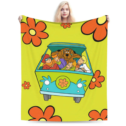 A person holds up a large blanket featuring a retro 1960s-style floral pattern and a cartoon van with characters inside, shown against a plain white background.