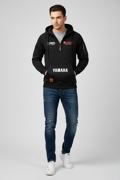A man modeling a black Yamaha hoodie with a front kangaroo pocket and white logo print, paired with blue jeans and white sneakers, against a light gray studio background.