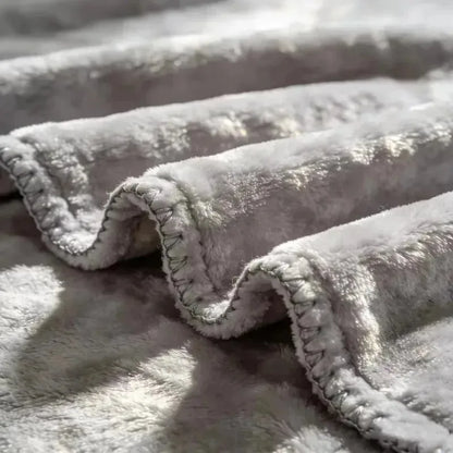 Close-up of a plush, soft gray blanket with a textured sherpa-like surface and stitched wavy edges, draped in folds with natural light and shadow.