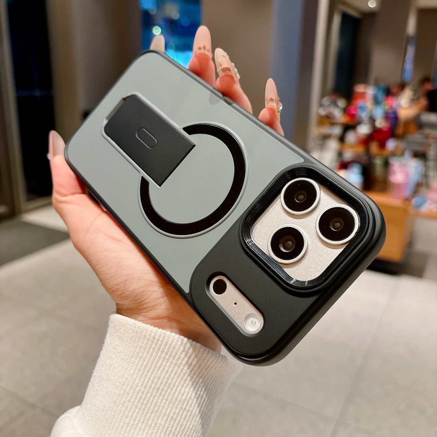 A hand holding a gray and black magnetic phone case with a built-in kickstand and camera cutout, shown in a blurred indoor retail environment.