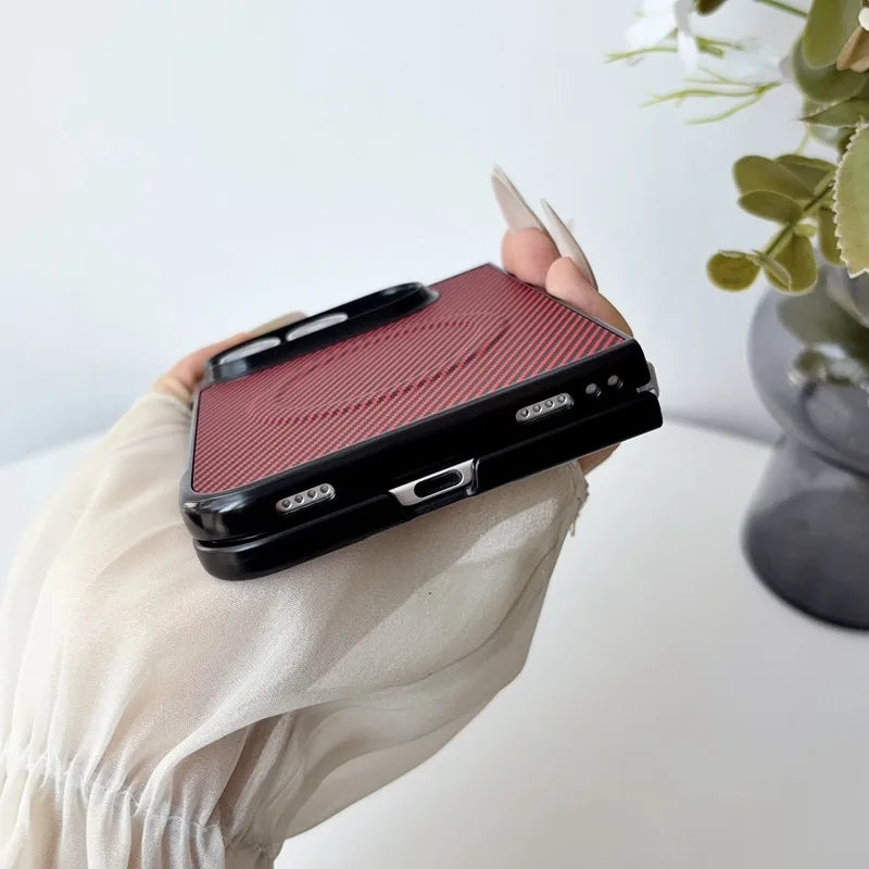 A hand wearing a cream-colored sleeve holds a black phone case with a textured red backplate featuring a circular magnetic design, set against a bright, softly blurred background with a plant in a vase.