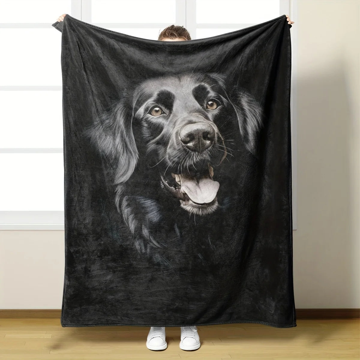 A person holds up a large black fleece blanket featuring a detailed grayscale portrait of a dog, shown in a home setting with a window and wooden floor.