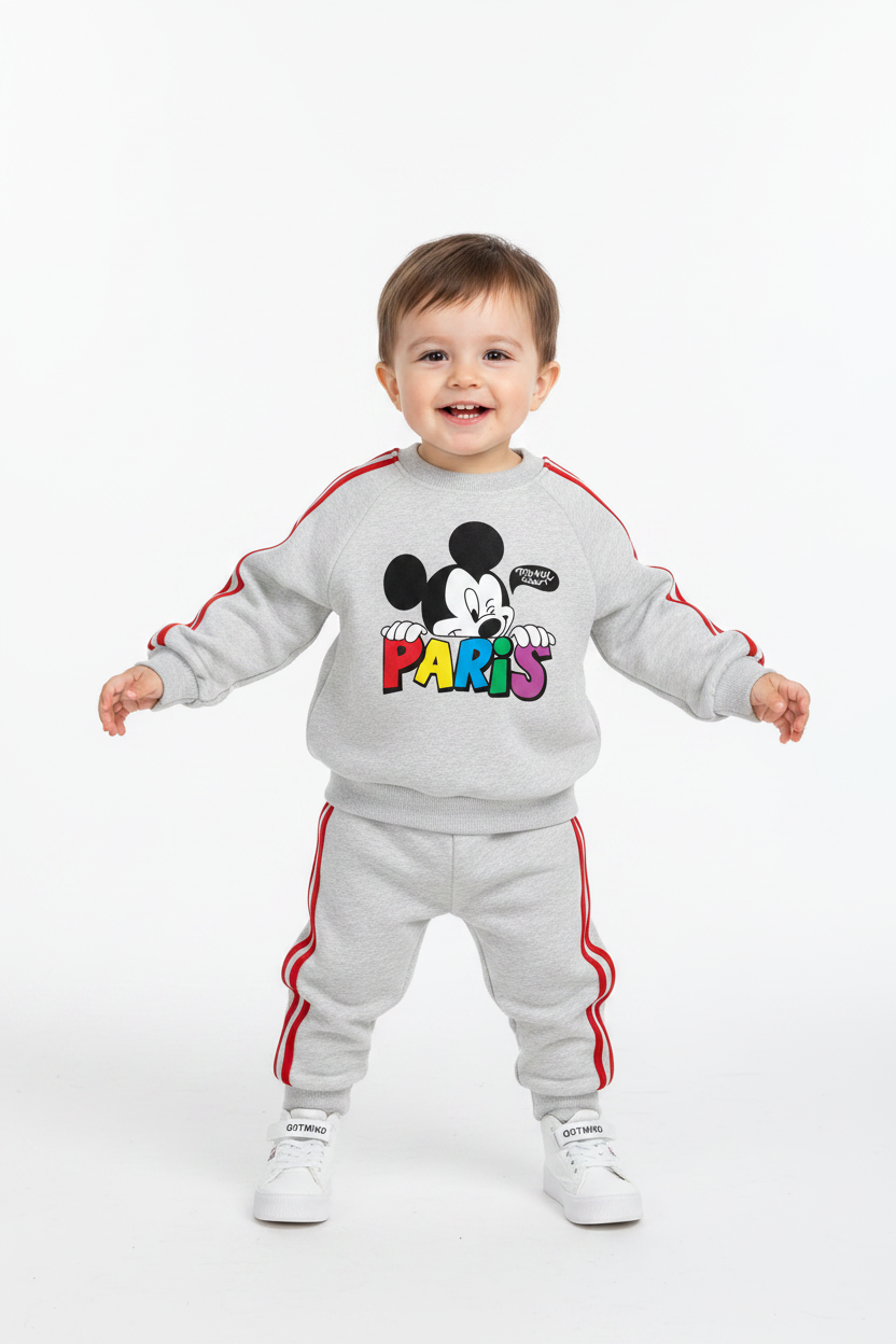 A toddler stands against a plain white studio background wearing a light gray sweatshirt and matching sweatpants set with red side stripes and a colorful Mickey Mouse 'PARIS' graphic on the chest. The child is also wearing white sneakers.