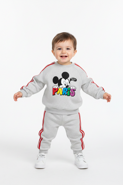 A toddler stands against a plain white studio background wearing a light gray sweatshirt and matching sweatpants set with red side stripes and a colorful Mickey Mouse 'PARIS' graphic on the chest. The child is also wearing white sneakers.