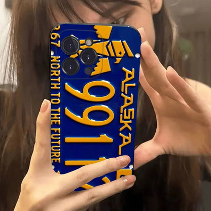 A person holding a blue phone case designed like an Alaska license plate with yellow numbers and text, photographed in a mirror selfie.