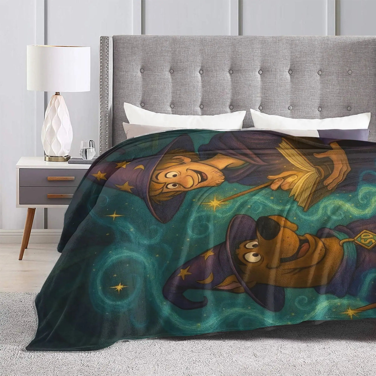A cozy bedroom scene with a bed made up with a dark teal blanket featuring a whimsical cartoon illustration of a wizard and a brown dog character. The room includes a gray tufted headboard, a white bedside table with a lamp, and a light gray rug.
