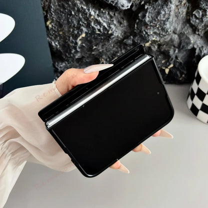 A black flip phone case with a slim black screen protector, held by a hand with long nude manicured nails. The background features a dark textured rock wall and a light gray surface with a black-and-white checkered cup.