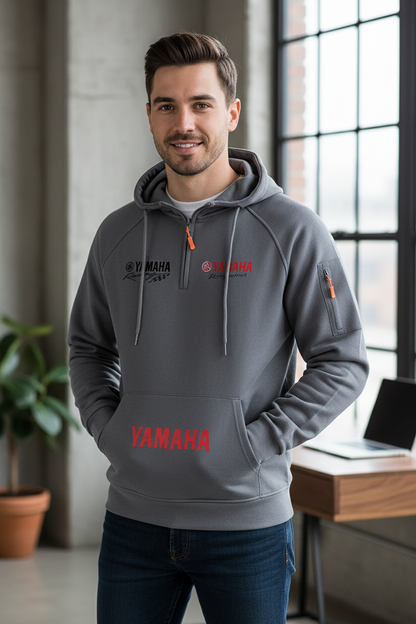 A man wearing a gray Yamaha hoodie with red and white graphics, standing in a modern indoor setting with a window, desk, and plant in the background.