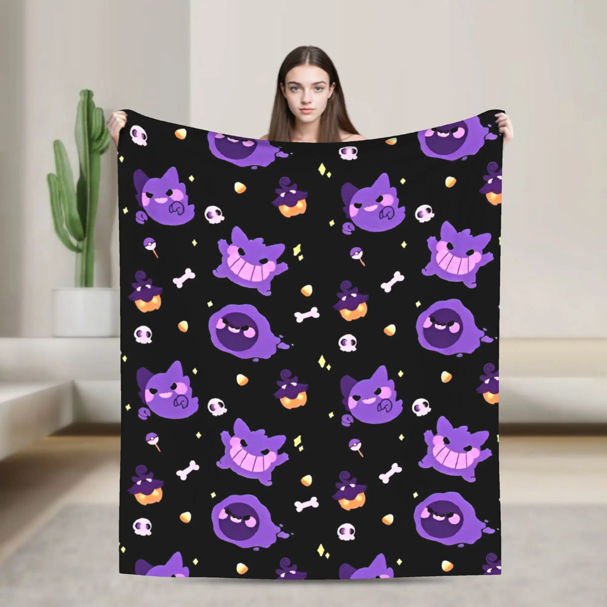 A woman holds up a black blanket featuring a repeating pattern of purple cartoon ghost-like characters, orange pumpkins, white skulls, and small bones. The background is a softly blurred indoor room with a cactus and light-colored walls.