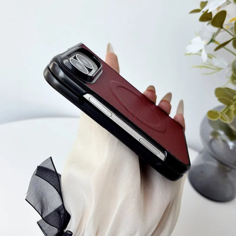 A hand holding a phone case with a textured burgundy back and black bumper edges, styled with a cream satin sleeve and a black ribbon bow, with a blurred vase of white flowers in the background.