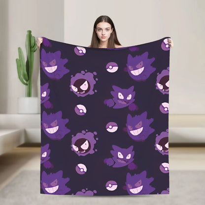 A woman holds up a large blanket featuring a repeating pattern of purple Gengar characters and Pokéball icons on a dark background, set in a modern living room with a cactus in the background.