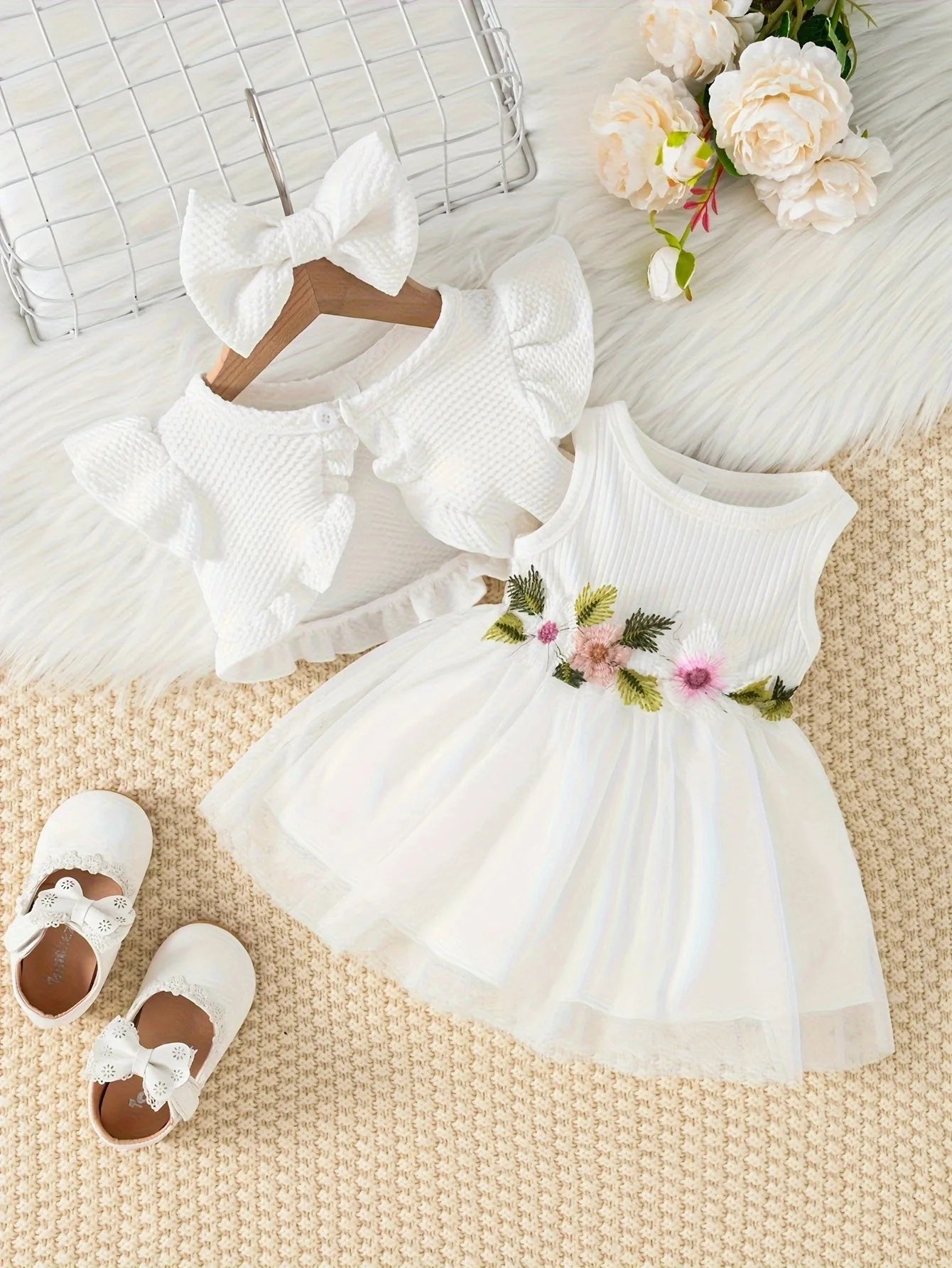 White baby dress with floral details, white shoes, and a white headband on a textured surface with flowers.