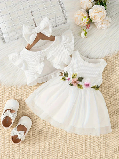 White baby dress with floral details, white shoes, and a white headband on a textured surface with flowers.