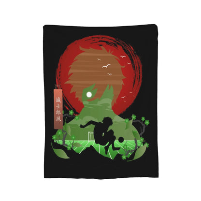 A black rectangular textile item, likely a towel or blanket, featuring a stylized graphic print of a soccer player in front of a red sun and green hills, with a small vertical brown tag containing white characters.