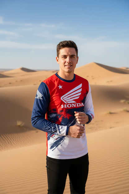 A man standing in a desert dune landscape wearing a red, white, and blue long-sleeve Honda racing shirt and black pants.