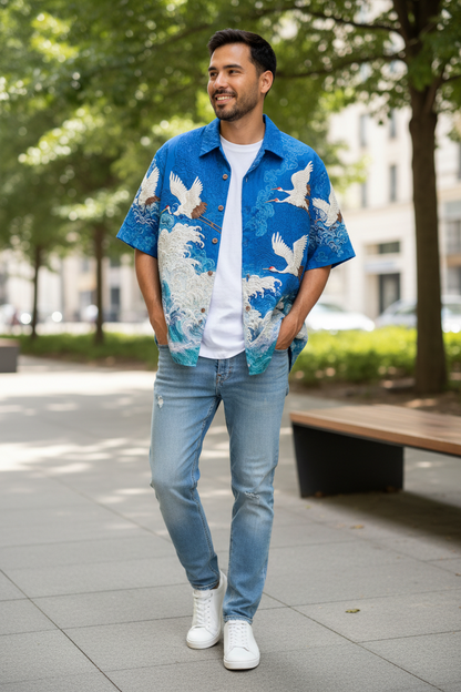 A man wearing a blue short-sleeve button-up shirt with a white and blue crane pattern, a white t-shirt, light-wash blue jeans, and white sneakers, walking on a paved outdoor path with trees in the background.