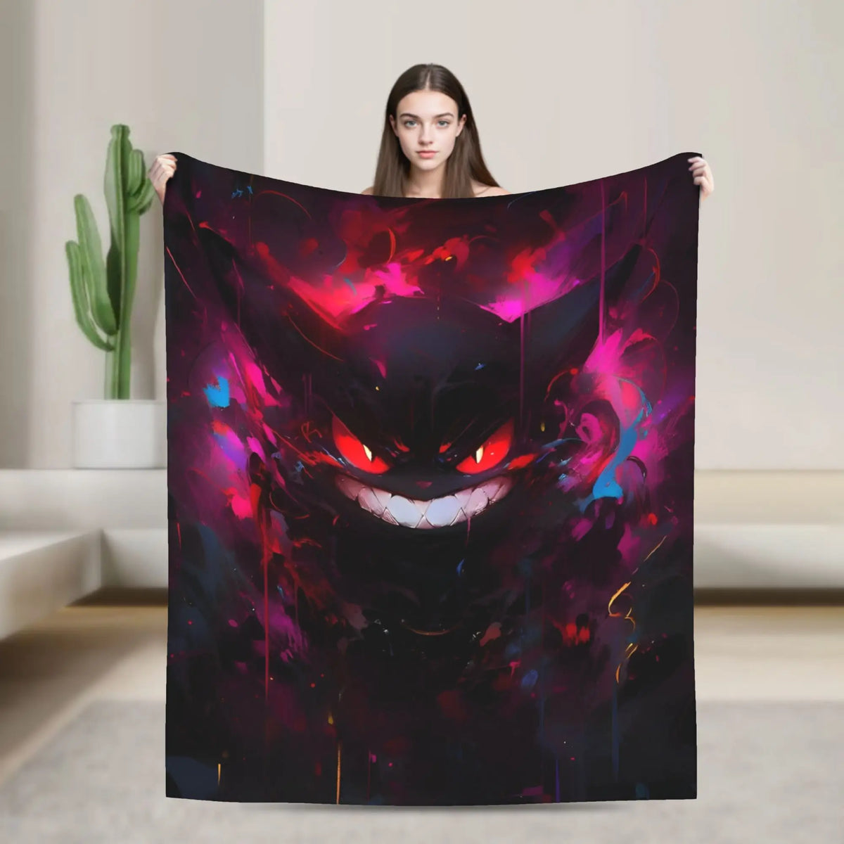 A woman holds up a large square blanket featuring a colorful, dark fantasy-style illustration of a grinning cat-like face with glowing red eyes and vivid pink, purple, and blue paint splashes on a black background.