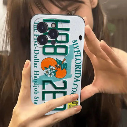 A close-up of a person holding a phone case featuring a Florida license plate design with orange graphics and text, photographed in a mirror selfie style.