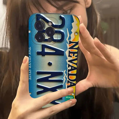 A person holding a phone case shaped like a vintage Nevada license plate, featuring raised blue numbers, yellow and white mountain graphics, and the words 'NEVADA' and 'TRIP SHAPED STATE'.