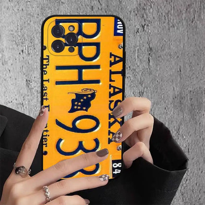 A phone case designed to look like an Alaska license plate, featuring a bright yellow background, dark blue lettering, and a small state flag graphic. The case is held by hands with silver rings and glittery nail polish against a gray concrete wall.