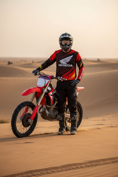 A person in a black, red, and yellow Honda motocross jersey and helmet stands on sand dunes next to a red Honda dirt bike.