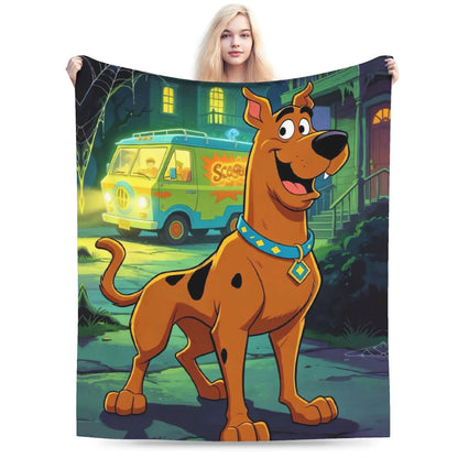 A person holds up a large printed blanket or tapestry featuring Scooby-Doo standing in front of a haunted house with the classic green van in the background. The blanket is shown against a plain white studio background.