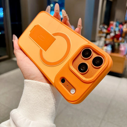 A hand with long manicured nails holds a bright orange phone case featuring a built-in kickstand and a textured metallic orange camera surround, set against a blurred indoor background.