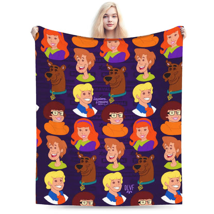 A person holds up a large blanket or throw featuring a repeating cartoon pattern of Scooby-Doo characters on a dark purple background. The blanket is shown against a plain white studio background.
