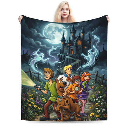 A person holds up a large printed blanket or tapestry featuring a Scooby-Doo night scene with the characters standing in front of a haunted house, ghosts, flowers, and a full moon. The product is shown against a plain white background.