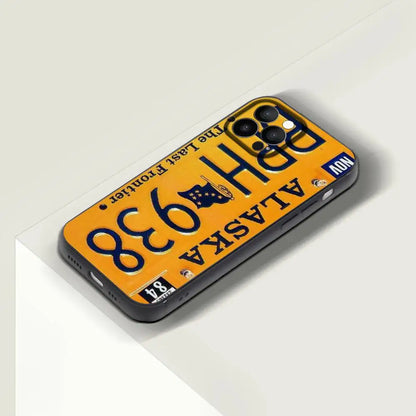 A phone case designed to look like an old Alaska license plate, featuring a yellow background with dark blue text and graphics, shown on a light gray angled surface.