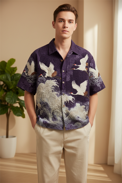 A man wearing a short-sleeve button-up shirt with a purple, white, and gray crane and wave pattern, paired with beige trousers, standing in a warm-toned indoor setting.