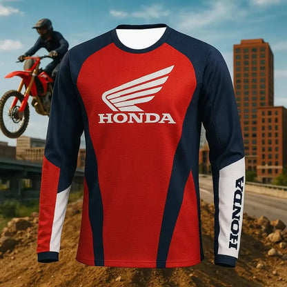 A long-sleeve Honda motocross jersey with a red and navy blue color-blocked design, featuring a large white Honda wing logo on the chest and 'HONDA' printed vertically on the sleeve. The jersey is shown against a blurred background of a dirt bike rider on a motorcycle and city buildings.