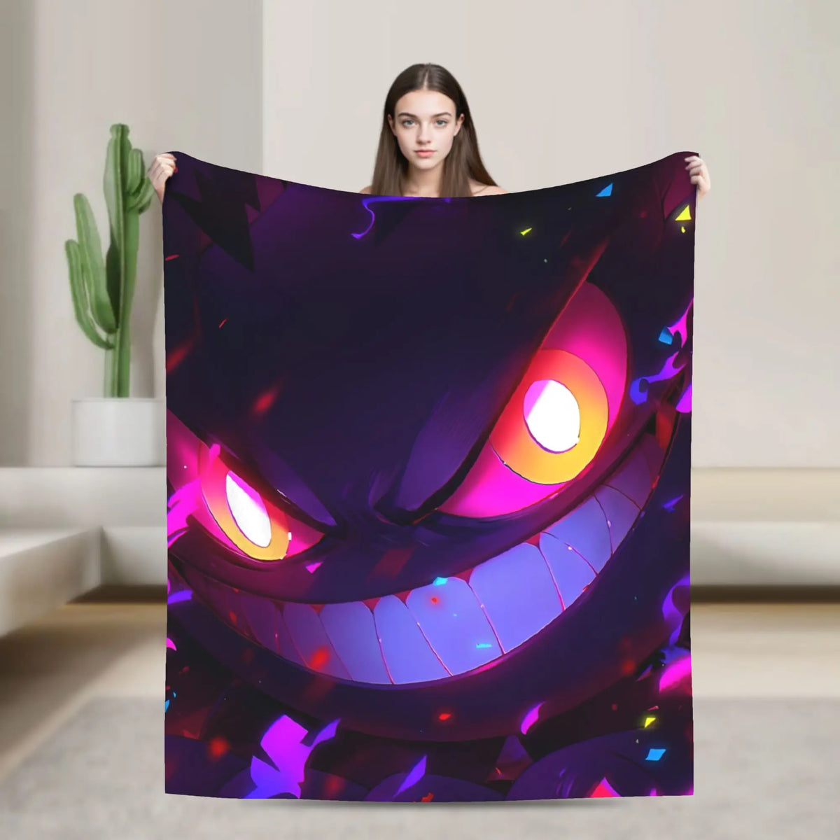 A woman holds up a large blanket featuring a colorful, stylized cartoon monster face with glowing pink and orange eyes and a wide grin. The blanket is shown in a brightly lit, modern room with a cactus in the background.
