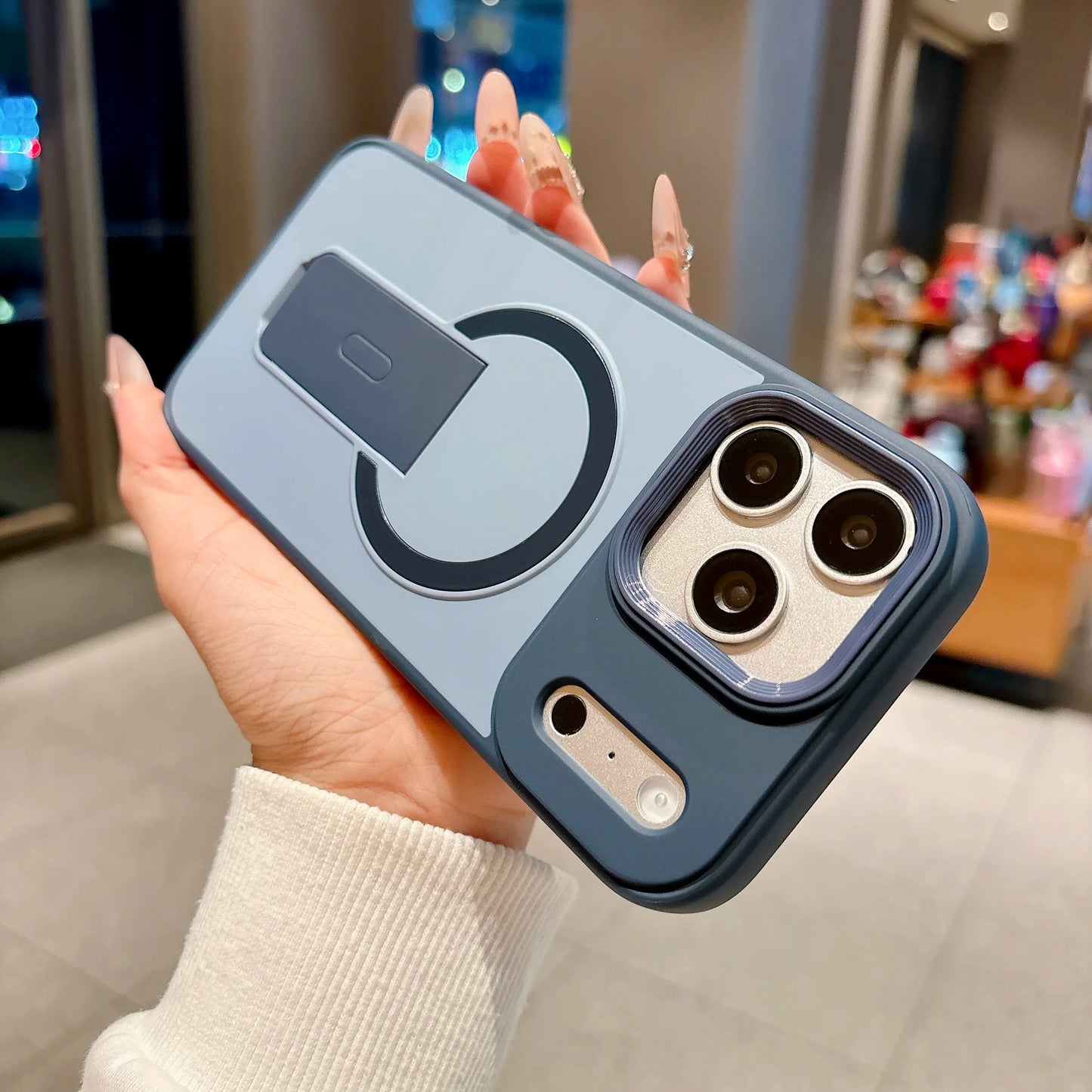 A hand holding a dark blue protective phone case with a built-in kickstand and circular magnetic ring, shown in a blurred indoor retail environment.