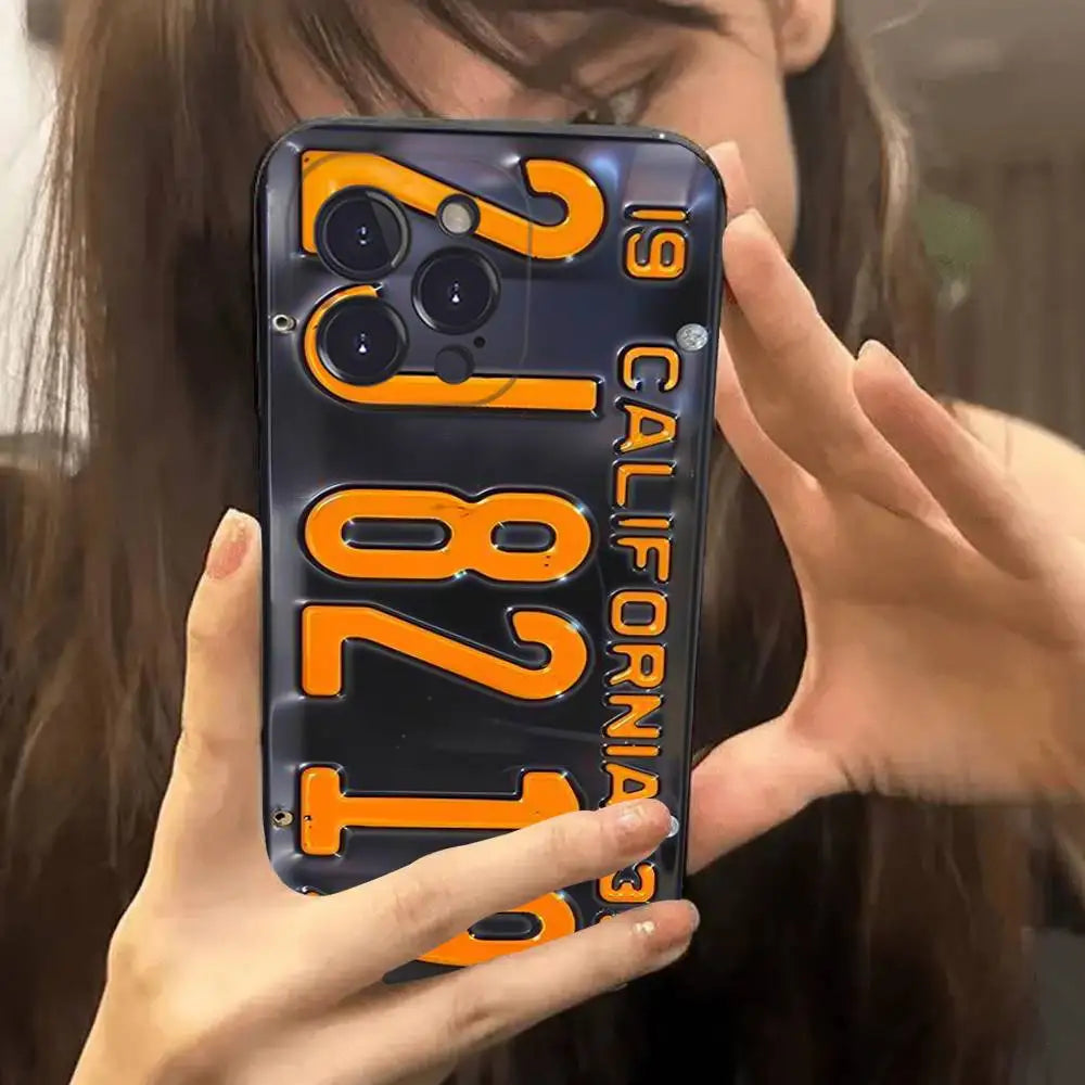 A close-up mirror selfie of a person holding a phone case featuring a vintage-style California license plate design with orange numerals and text on a dark background.