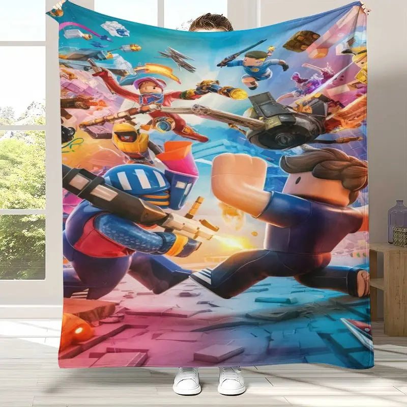 A person holds up a large fleece blanket featuring a colorful, dynamic cartoon battle scene with block-style characters, vehicles, and bright action effects. The blanket is shown in a home setting with a window and floor visible in the background.
